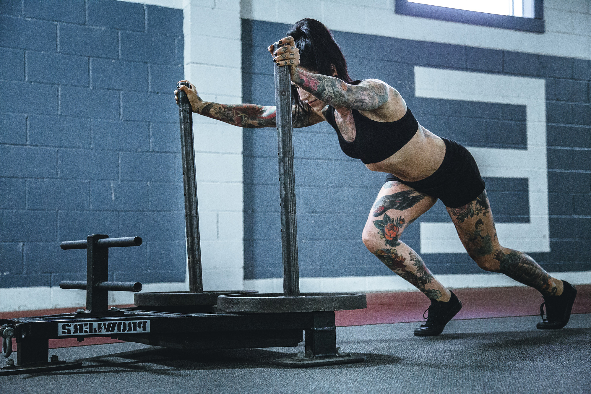 A tattooed athlete pushes a weighted sled across a gym. As a crossfit coach she can focus on her work because she has crossfit trainer insurance.