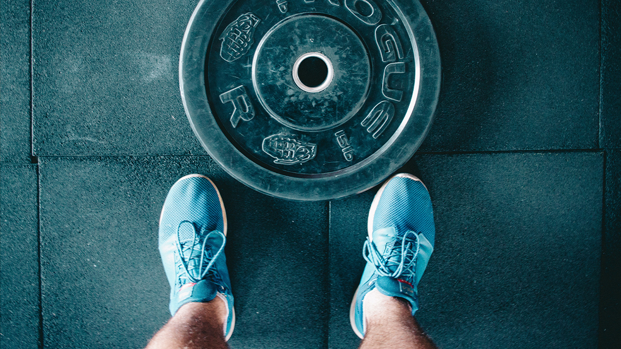 From above, feet in gym shoes stand in front of a plate weight. This gym's trainer is covered with fitness instructor insurance.